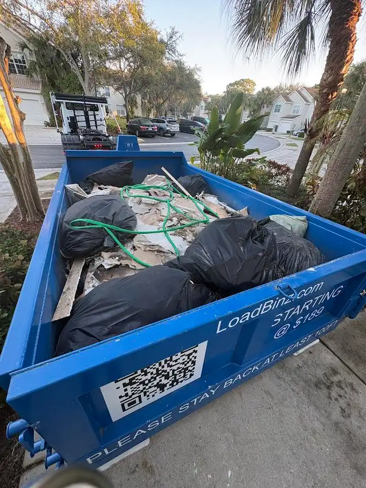 A blue LoadBinz dumpster filled with residential waste bags and debris during a garage cleanout project.