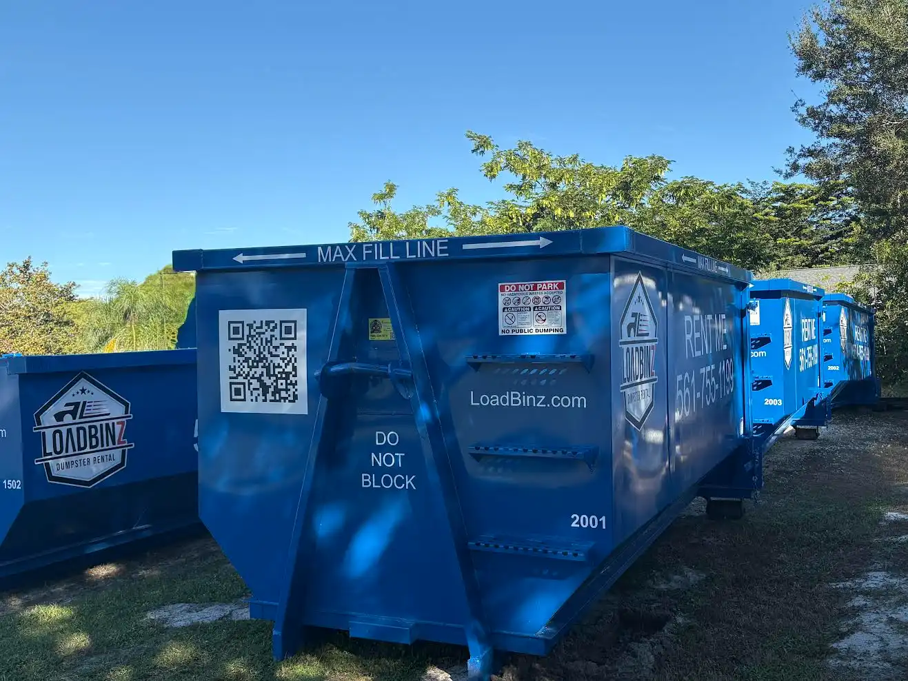 A row of blue LoadBinz rental dumpsters featuring 'Max Fill Line' markers, QR codes, and contact information for local dumpster services.