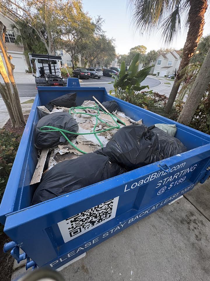 A blue LoadBinz dumpster filled with residential waste bags and debris during a garage cleanout project.