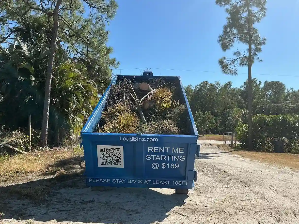 A blue LoadBinz roll-off dumpster filled with yard waste and tree trimmings, parked on a dirt lot with rental pricing and a QR code visible.