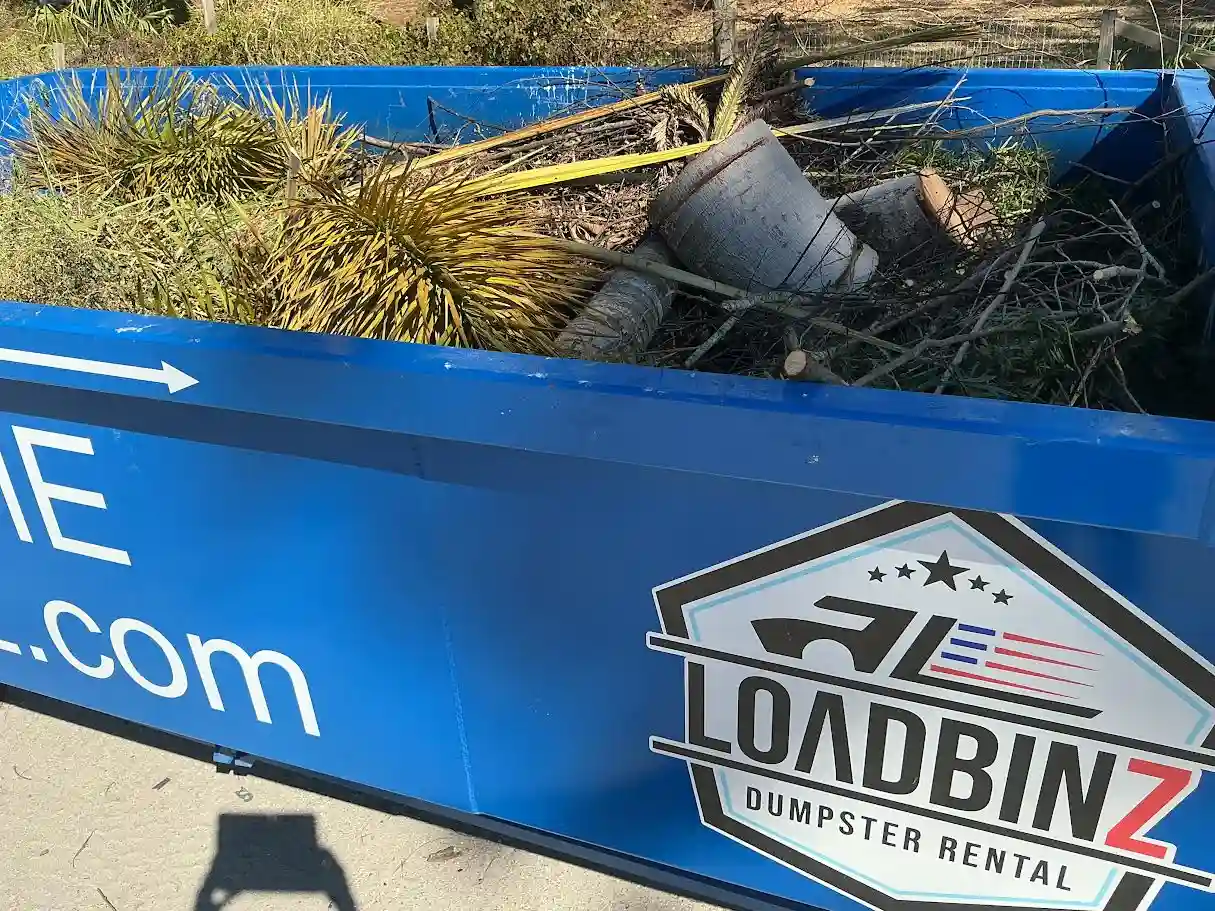 A close-up view of a blue LoadBinz dumpster filled with cut palm fronds, branches, and tree trunks, highlighting the company logo.