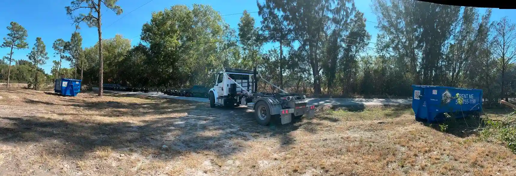A white roll-off dumpster truck parked on a dirt lot in a rural setting, positioned between two blue LoadBinz dumpsters.