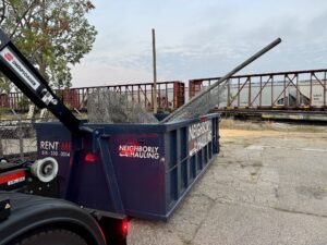 Neighborly Hauling hook-lift dumpster loaded with fencing material and metal poles near a rail yard.