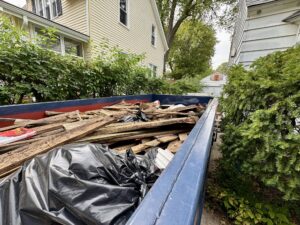 Dumpster filled with construction debris, including old wood and trash bags, placed between two houses.